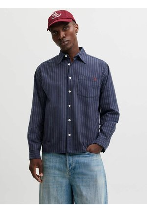 Homme portant une casquette rouge et une chemise bleue à fines rayures avec poche, debout avec une main dans la poche d'un jean bleu clair, sur un fond uni.