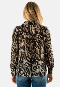 Blusa transparente en beige y negro con estampado animal, con cuello, mangas largas y detalles de botones. La parte trasera presenta un pliegue para mayor movilidad.