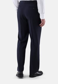 Mann trägt dunkelblaue Anzughose, schwarze Lederschuhe und ein weißes Langarmhemd, von hinten vor einem einfarbigen Hintergrund gezeigt.