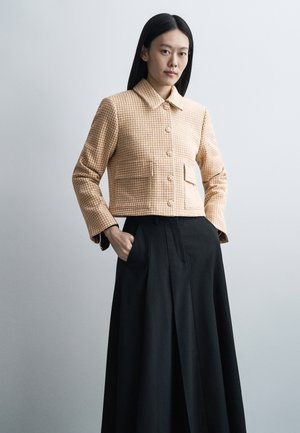 Femme aux longs cheveux noirs portant une veste beige à carreaux boutonnée et une longue jupe noire, debout, les mains dans les poches, devant un fond uni.