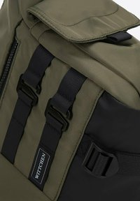 Zielony plecak z tkaniny z czarnymi akcentami, regulowanymi paskami, zakładką oraz oznaczoną metką. Powierzchnia ma gładką teksturę z detalami przeszyć.