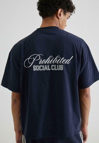 Tamsiai mėlynas medvilninis marškinėliai su užrašu "Draudžiamas socialinis klubas" baltos ir sidabrinės spalvos ant nugaros, su laisvu, atsipalaidavusiu siluetu.