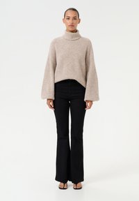 Maglione beige a collo alto lavorato a maglia con maniche ampie, abbinato a jeans neri a vita alta e a zeppe nere, su uno sfondo neutro.