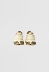 Beige sneakers with a textured upper and a cushioned heel, featuring a clear rubber sole with a brown accent at the base and logo detail.
