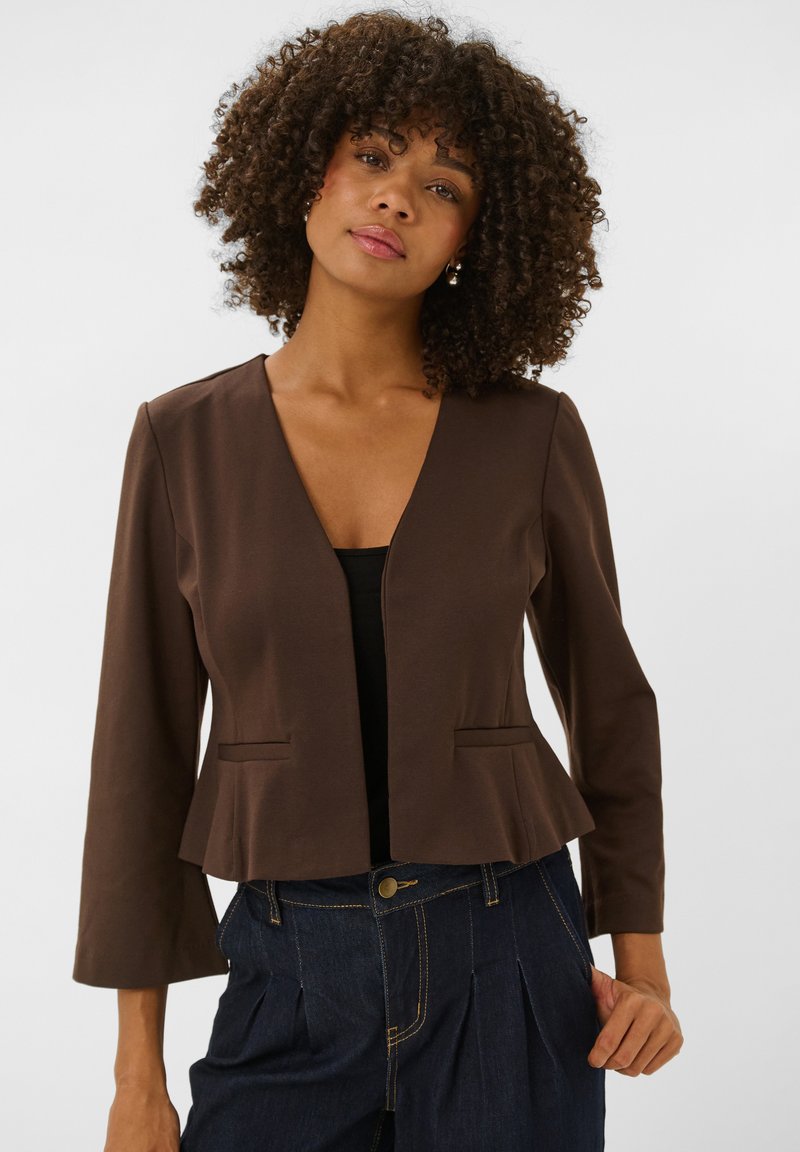 Femme aux cheveux bouclés portant un blazer marron ouvert sur un top noir et un jean plissé foncé, sur un fond uni.