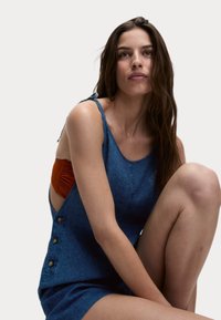 Vestido de mezclilla con un cuerpo ajustado, tirantes anchos y lados abiertos. Presenta tres botones decorativos en el lado izquierdo. Color azul, textura suave.