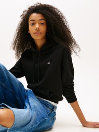 Svart hettegensere med kengurulomme, brukt sammen med løse blå denimshorts med slitte detaljer. Modellen har krøllete hår og et nøytralt uttrykk.