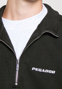 Černá zipová mikina s texturovanou tkaninou, žebrovým límcem a bílým logem "PEGADOR" na hrudi. Zip má stříbrné táhlo.