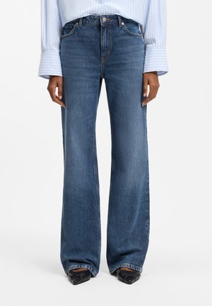 Person iført højtaljede, vidde-blå jeans kombineret med en lyseblå, stribet skjorte og sorte spidse sko.