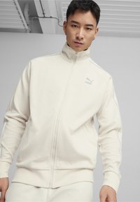 Vit zip-up jacka i texturerat material, med ståkrage och ljusblå accentband längs ärmarna. Logotyp på bröstet.