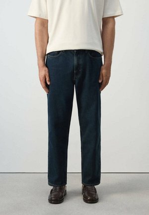 Hombre con vaqueros azul oscuro de corte recto, una camisa blanca de manga corta y mocasines marrón oscuro, de pie frente a un fondo liso.
