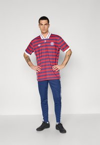 Czerwona i niebieska koszula z krótkim rękawem w wzory z białym kołnierzykiem, zestawiona z granatowymi spodniami z białymi paskami po bokach. Całość uzupełniają czarne sneakersy.