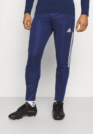 Homme portant un pantalon de survêtement Adidas bleu marine à bandes blanches, des chaussettes Adidas blanches et des chaussures de football Adidas Nemesis noires.