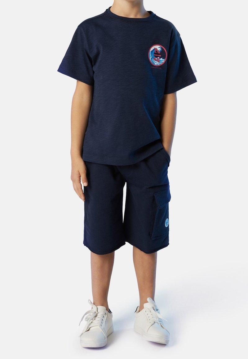 Enfant portant un t-shirt bleu marine avec un motif de vague et un short cargo assorti, debout avec une main dans la poche et des baskets blanches.