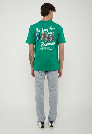 Uomo con maglietta verde con orsetti colorati e scritta bianca, pantaloni grigio chiaro e scarpe da ginnastica bianche, in piedi di spalle.