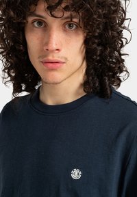 Młoda osoba z kręconymi, ciemnymi włosami, nosząca granatową koszulę z małym białym okrągłym logo na piersi, z neutralnym wyrazem twarzy.