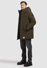 Olivengrøn pufferjakke med høj krave, lynlåslommer og logopatch. Samlet med mørkegrå jeans og sorte klumpede støvler.