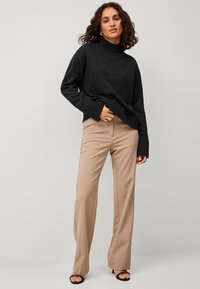 Schwarzer Strickpullover mit Rollkragen und gerippten Bündchen, kombiniert mit beigen, taillierten Hosen; die Schuhe sind offene schwarze High Heels.