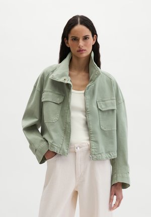 Kobieta o długich ciemnych włosach nosi jasnooliwkową krótką kurtkę na białym topie i jasnobeżowych spodniach z wysokim stanem, stojąc na jednolitym tle.