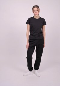 Svart bomull t-shirt och svarta träningsbyxor, båda med en subtil logotyp på bröstet och låret. Modellen bär vita sneakers. Enfärgad bakgrund.