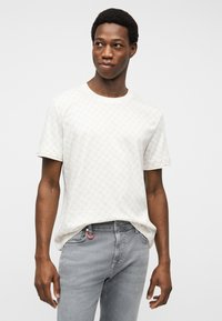 Νέος άνδρας που φοράει λευκό κοντομάνικο μπλουζάκι με σχέδιο και ανοιχτό γκρι τζιν, στέκεται με τα χέρια χαλαρά και κοιτάζει δεξιά.