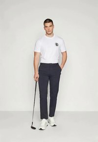 Jeune homme se tenant avec une main dans la poche, tenant un club de golf, portant un t-shirt blanc, un pantalon sombre et des baskets blanches sur un fond uni.