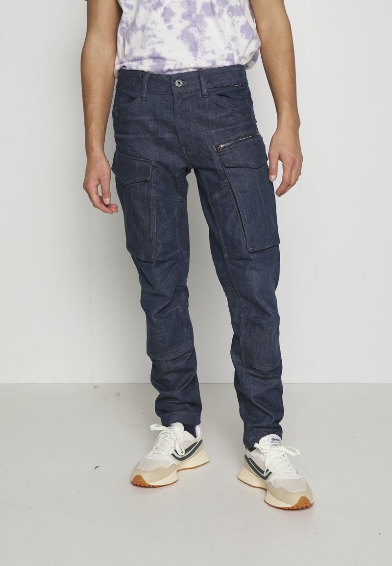 Jean cargo bleu marine avec plusieurs poches, détails zippés et une coupe légèrement fuselée. Confectionné en denim au toucher lisse.