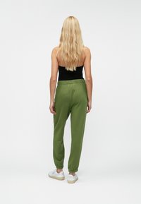 Green cotton joggers with an elastic waistband and cuffed ankles, paired with white sneakers. Back view, model wearing a black top.