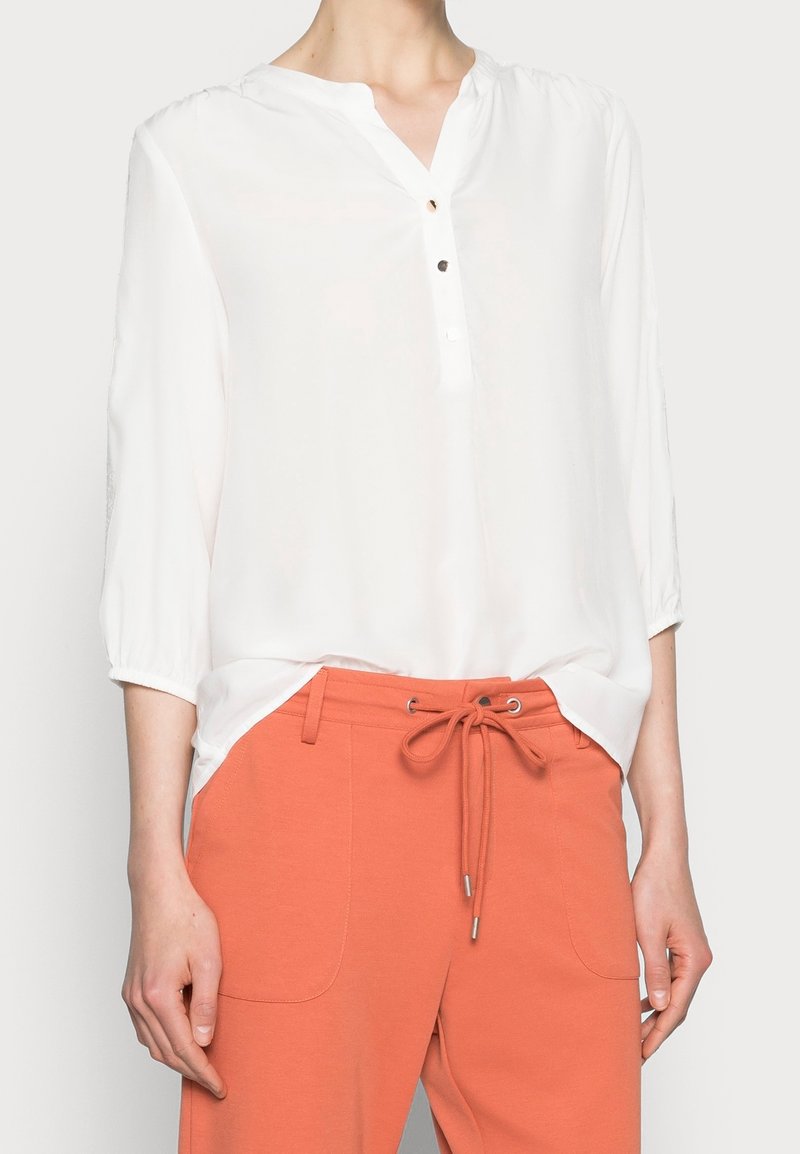 Vrouw die een losse witte blouse draagt met driekwart mouwen en koraalkleurige broek met trekkoord en voorzakken.