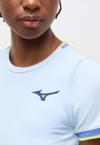 Κοντινή λήψη ενός ατόμου που φοράει ανοιχτό μπλε αθλητική μπλούζα με λογότυπο σε σκούρο μπλε στο στήθος και κίτρινη και μπλε διακόσμηση στον ώμο και το μανίκι.