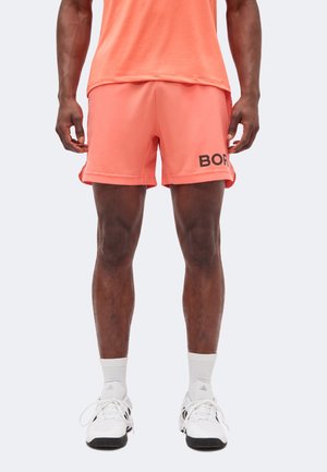 Homme portant un short de sport corail avec le texte noir "BOR" sur la jambe gauche, des chaussettes blanches et des chaussures de sport blanches Adidas, debout sur un fond clair.
