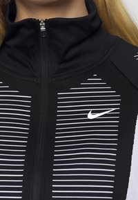 Chaqueta deportiva negra con cremallera y cuello alto, con franjas blancas horizontales y un pequeño logotipo blanco de Nike en el lado izquierdo.