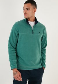 Pullover in pile verde con collo a zip, colletto scuro in contrasto e logo ricamato. Texture morbida e vestibilità rilassata.