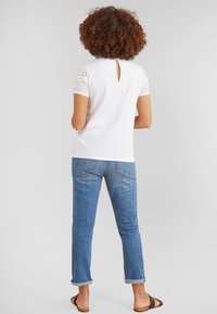 Haut blanc à manches courtes avec des accents en dentelle et un détail à l'arrière en forme de goutte, associé à un jean en denim bleu clair avec revers roulés et des sandales marron.