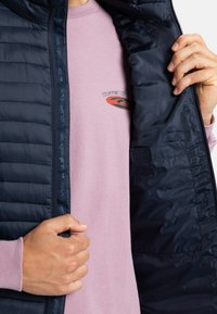 Hände ziehen eine marineblaue gesteppte Jacke auf und zeigen ein rosa Hemd mit einem kleinen runden "Tastes Like"-Logo auf der Brust.