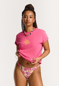 T-shirt di cotone rosa con maniche arrotolate, presenta testo dorato "Come Fanno i Locali." Indossato con un bikini a fantasia floreale nei toni del rosa e del verde.