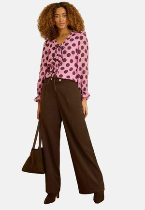 Femeie purtând o bluză roz cu buline negre mari, pantaloni maro cu picior larg, cizme maro cu toc și ținând o geantă maro.