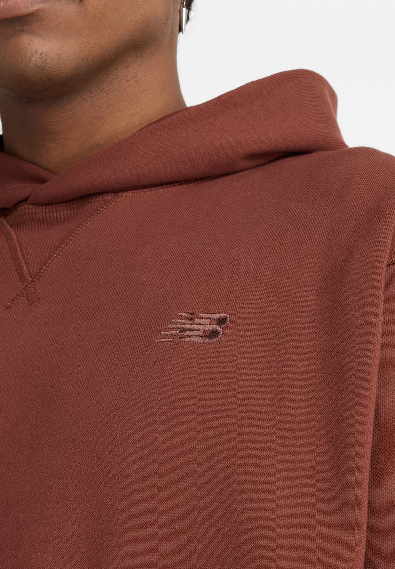 Maroon hooded sweatshirt made of soft fabric. Features a raised New Balance logo on the chest. Simple design with seamless edges.
