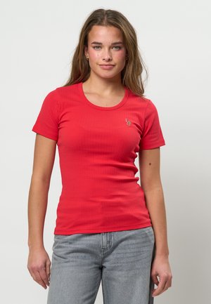 Mlada žena u uskoj crvenoj majici kratkih rukava i svijetlo sivim trapericama, stoji ispred jednobojne bijele pozadine.