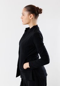 Maglietta nera a maniche lunghe con colletto alto e tasche. Presenta una texture liscia e un design aderente, con cuciture visibili lungo i lati.