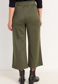 Olivegrüne Schlaghosen aus einem glatten Stoff mit einem elastischen Taillenbund, kombiniert mit schwarzen Knöchelstiefeln.