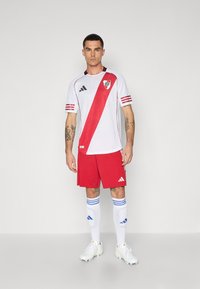 Fotbollströja i vitt med ett diagonalt rött streck, röda shorts, vita strumpor med blå ränder och vita fotbollsskor. Logotyp på bröstet.