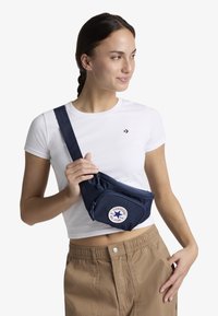 Marsupio blu navy realizzato in tessuto resistente, dotato di tasca frontale con zip e patch circolare del logo Converse. Cinghia regolabile per il massimo comfort.