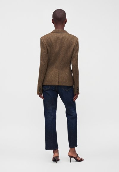 Personne aux cheveux courts portant un blazer marron, un jean bleu foncé et des sandales ouvertes à talons marron, debout de dos sur un fond neutre.