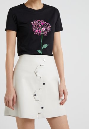 Frau trägt ein schwarzes T-Shirt mit einem pinkfarbenen Blumenmotiv und einen weißen Rock mit Zickzack-Knopfdetails an der Vorderseite.