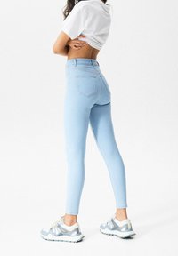 Jeans de tiro alto de color azul claro con un corte ajustado, cinco bolsillos y una textura de denim suave. Combinados con zapatillas deportivas de color claro.