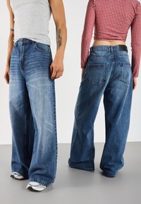 Weite blaue Denim-Jeans mit einem ausgewaschenen Finish; enthalten Taschen und Gürtelschlaufen. Getragen von zwei Personen in lässiger Kleidung.