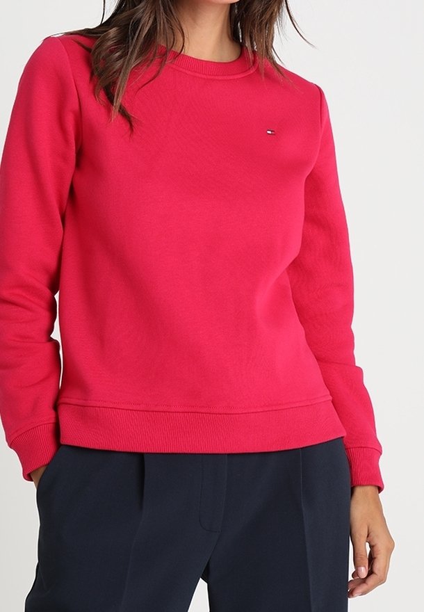Femme portant un pull rouge vif à manches longues et col rond, associé à un pantalon bleu marine foncé, debout avec une main dans la poche.