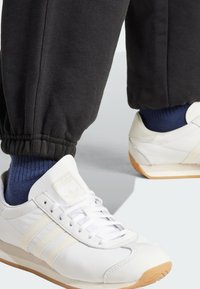 Bílé sportovní boty s třemi světle šedými pruhy, mají zaoblenou špičku a textured podrážku. V kombinaci s černými zkrácenými tepláky a modrými ponožkami.