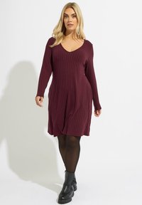 Robe bordeaux côtelée à manches longues, décolleté en V et ourlet évasé, associée à des bottines noires et des collants noirs transparents.